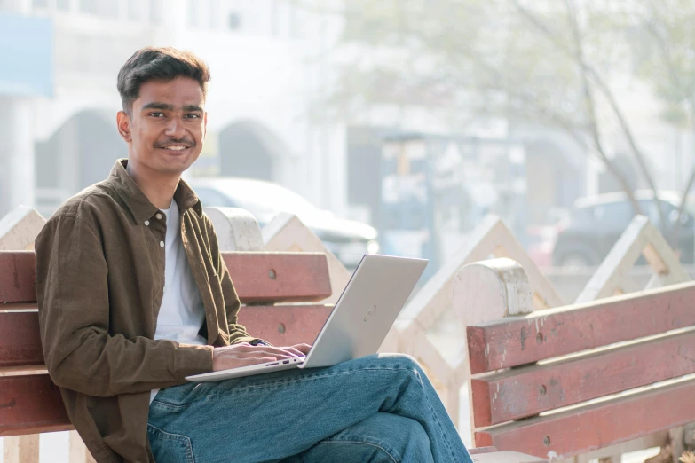 a young man sits on a park bench and uses his laptop