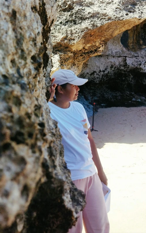 a  in pink sweatpants and a white hat is standing in front of a rock formation and looking at the water