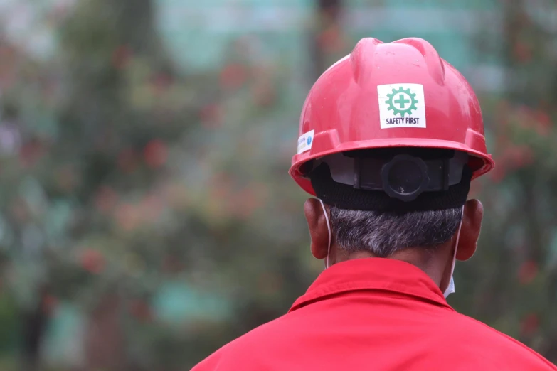 the back view of a man with a hard hat, red jacket and some trees in the background