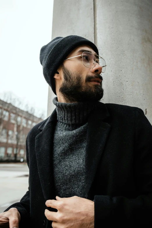 a man wearing glasses and a beanie leaning on a wall