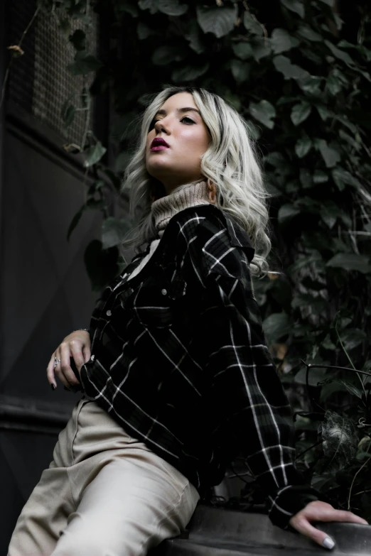 blonde woman posing on a cement base near a leafy wall