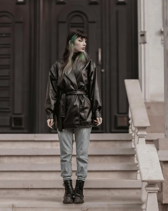 a person in a black leather coat and blue jeans stands in front of stairs with green hair