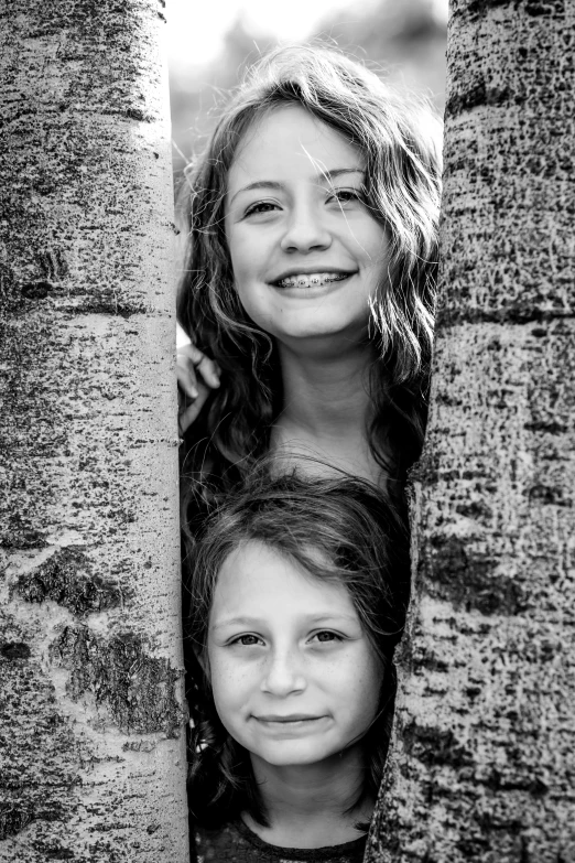 a couple of young ladies peeking out from behind a tree