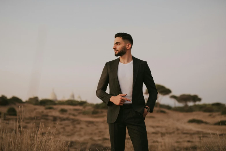 man in black suit standing near dry grass with one hand on his hips