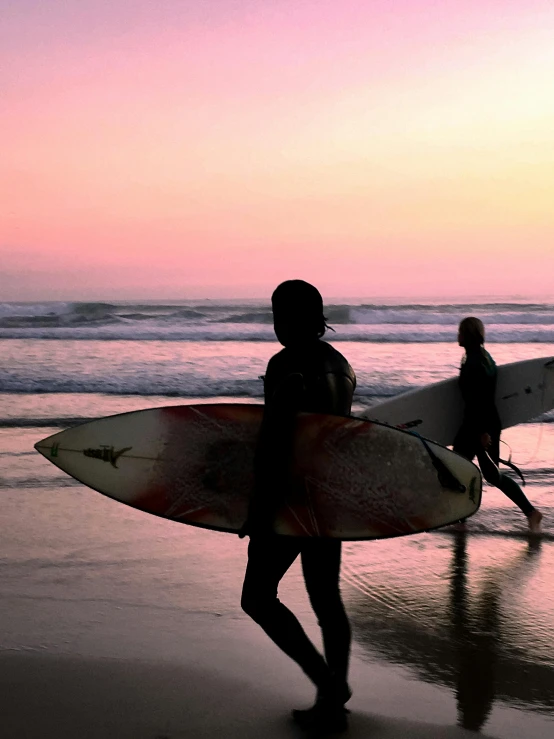 two people are on the beach walking with their surfboards