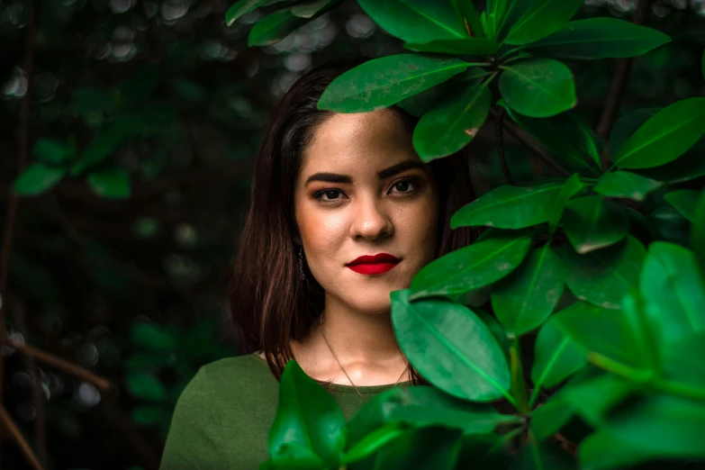 a woman standing behind green leaves wearing a red lipstick