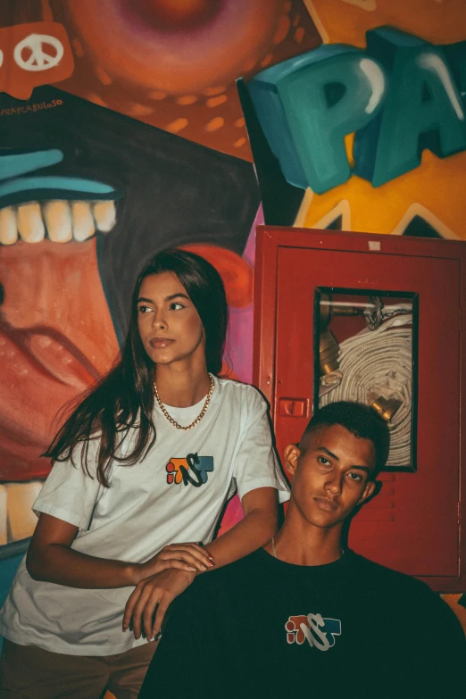 two young people sitting down together in front of a wall with various colorful paintings