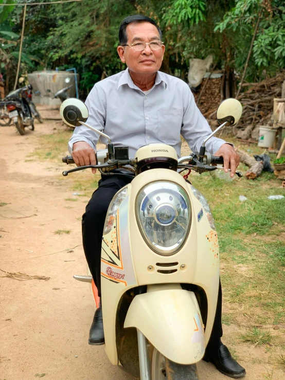 an older man with glasses sitting on a moped in front of a small group of people