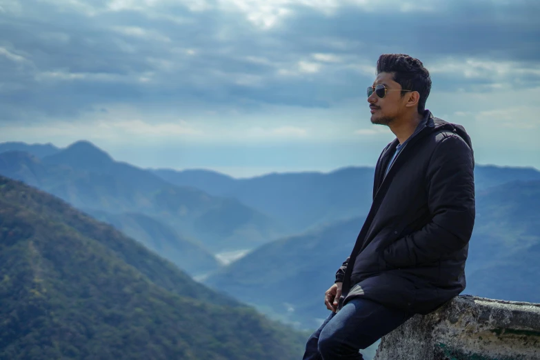 a man wearing a coat and glasses sitting on top of a mountain