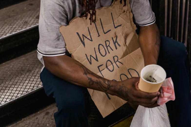 a man sits on a step holding a sign and drinking a cup of coffee