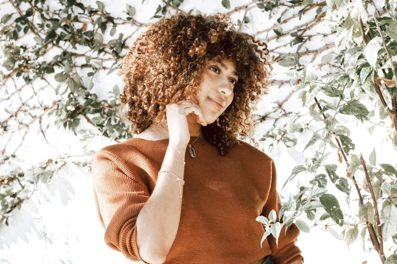 a young woman with curly hair, in a brown sweater, stands in front of a tree