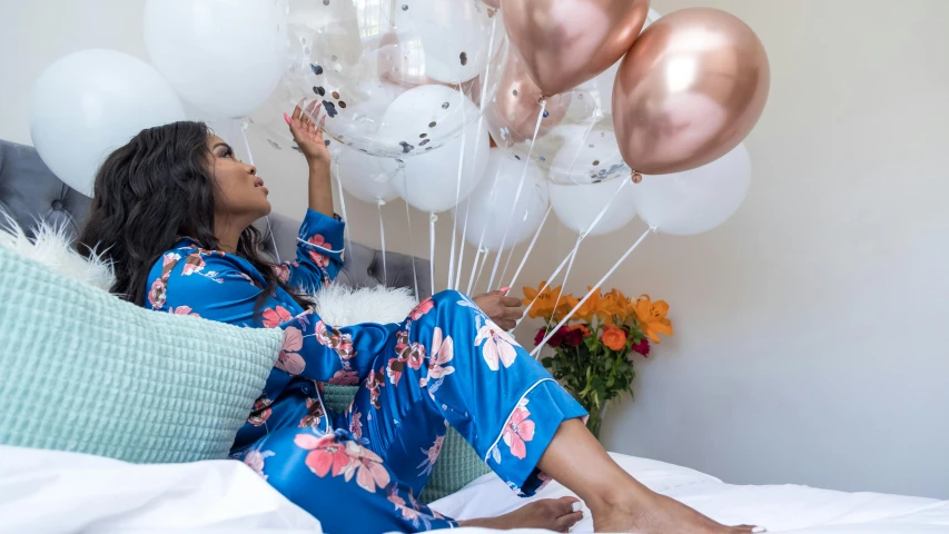 an attractive black woman in a blue floral pajamas is holding a bunch of white and gold balloons