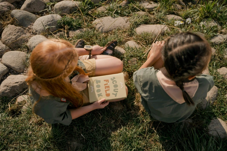 two girls sitting on the ground looking up at the sky with a book