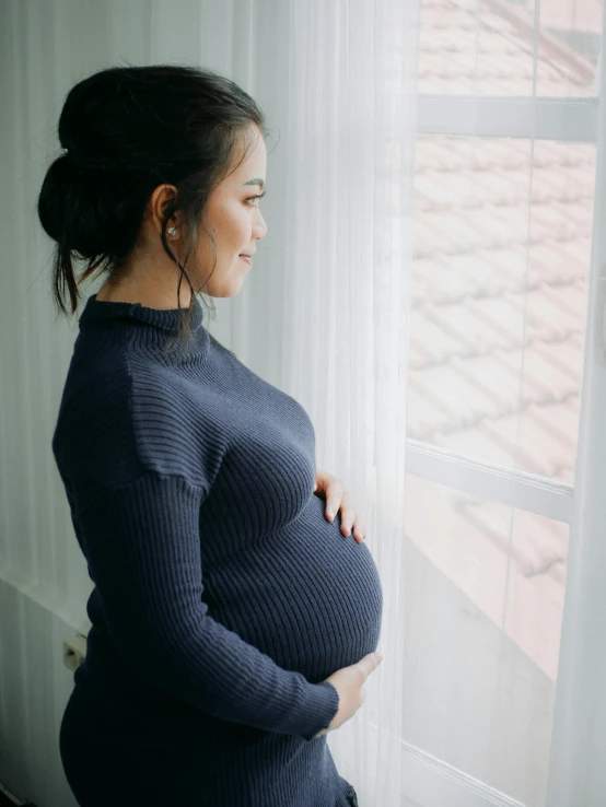 a pregnant woman stands next to a window