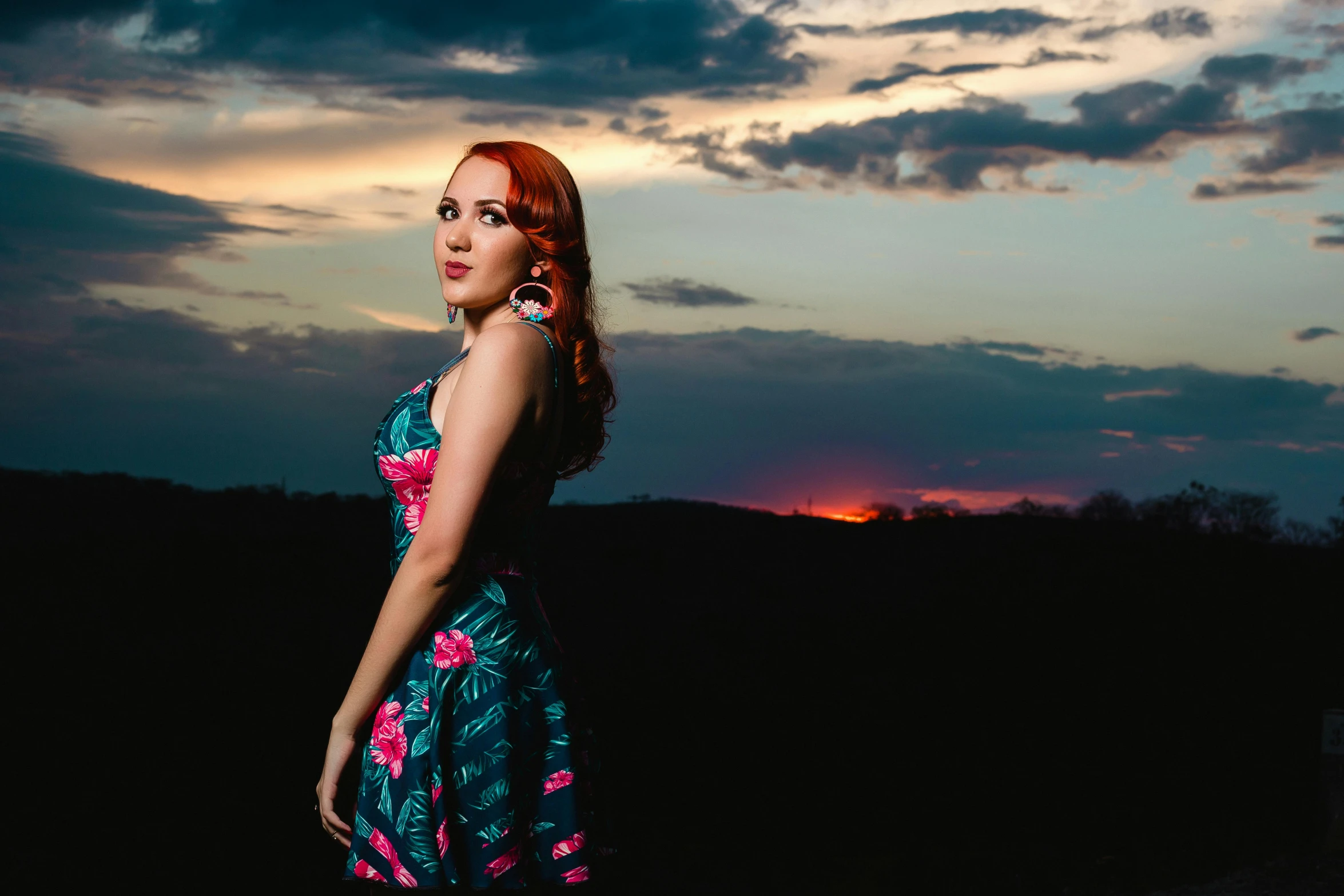 a woman in a dress with floral print poses for a po in front of the sunset