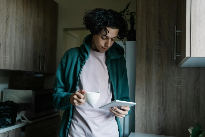 a young man stands in the kitchen holding a tablet and drinking coffee