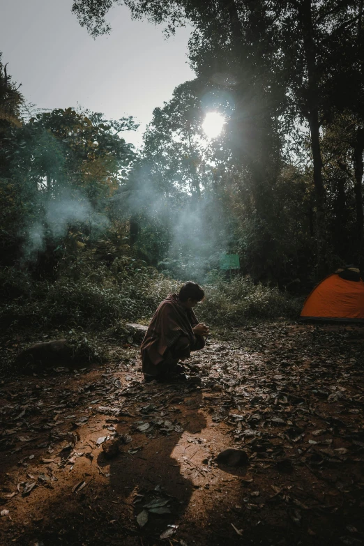 man crouching down in a forest next to his tent