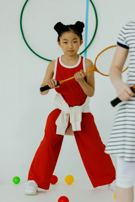 girl in red jumpsuit holding tennis racket next to a white wall