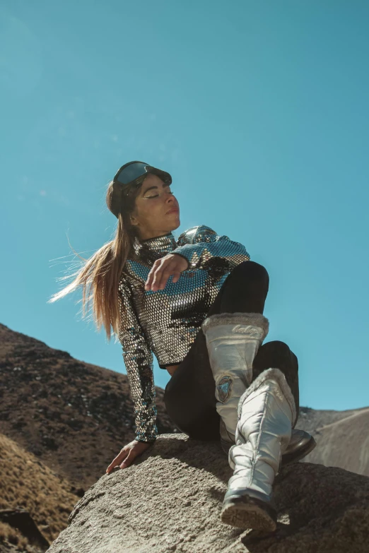 a woman in silver jacket and cap sitting on a rock