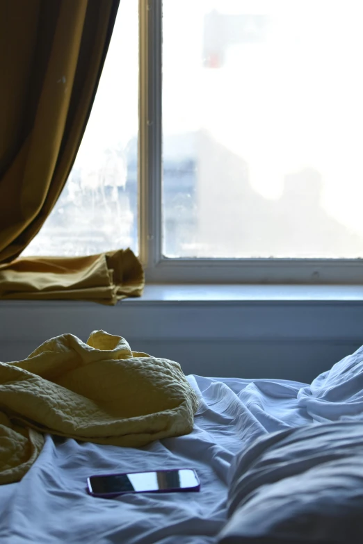 a phone laying on a unmade bed next to the window