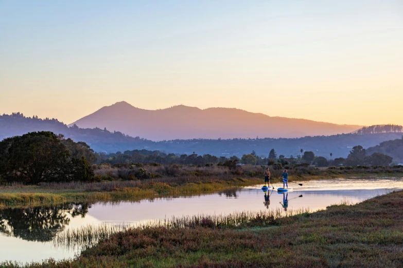 two people holding surf boards standing in a lake with mountains in the background