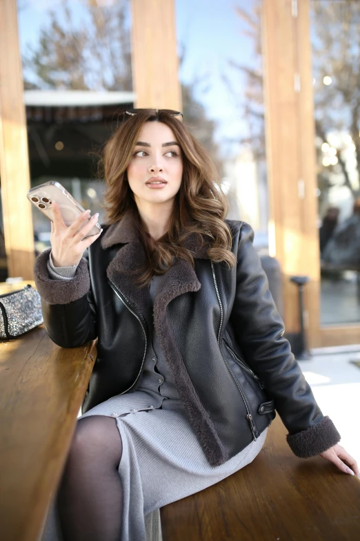 a woman in a leather jacket sits at a table and holds a cellphone