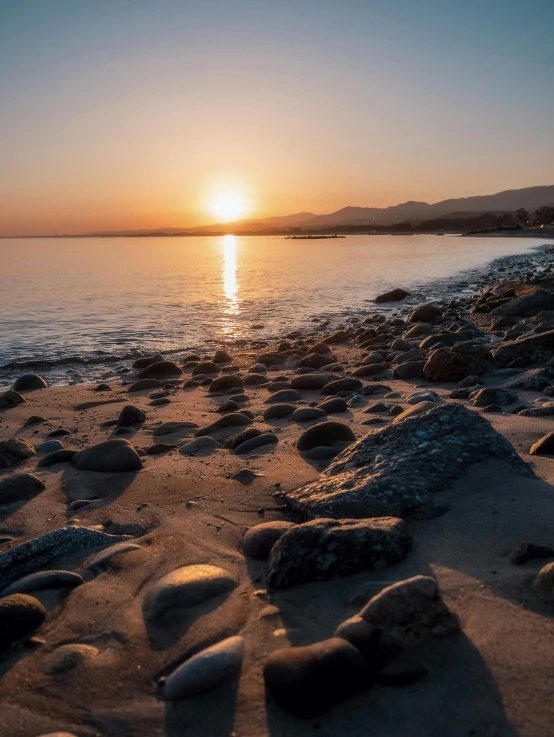 the sun sets over a large body of water with rocks and sand
