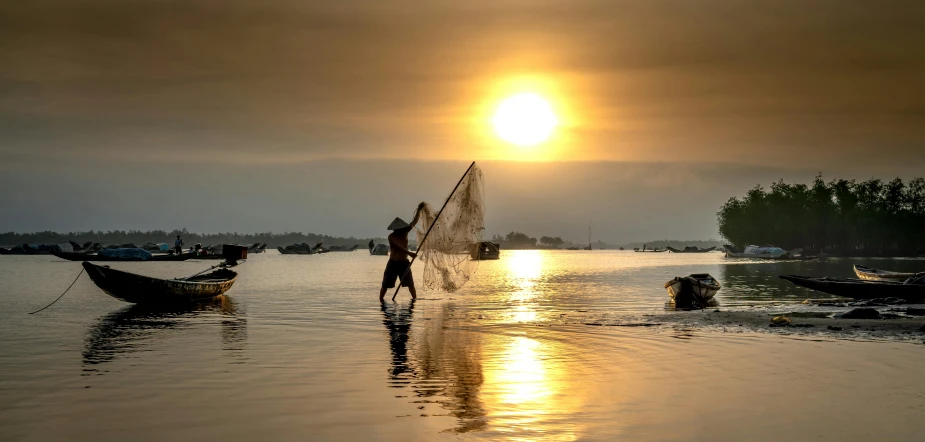 a man standing in the water, holding a net while setting sun over boats