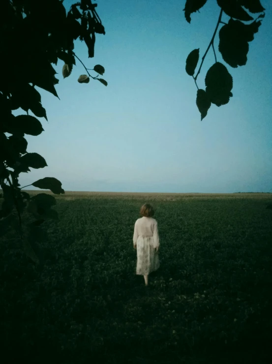 a girl wearing white stands alone in the middle of a green field