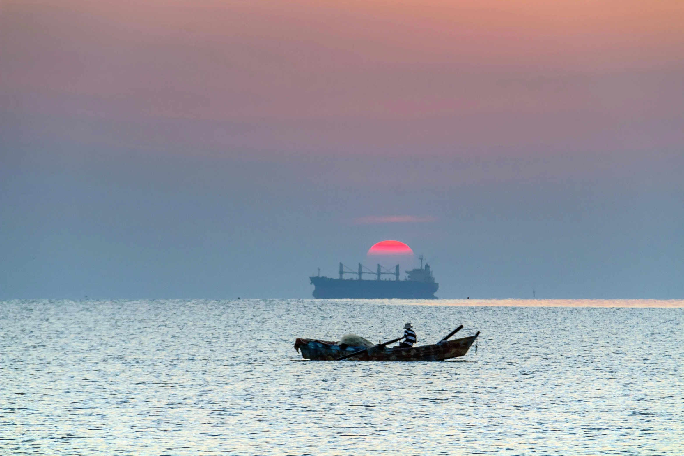 boat out on the ocean at dusk with a large cargo ship in background