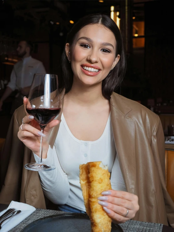 a smiling woman poses for the camera while holding up a glass of wine and a pastry