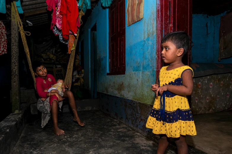 a little girl in a yellow dress stands near two other children sitting and standing outside