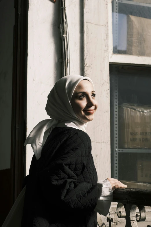 a woman wearing a headscarf stands by a window