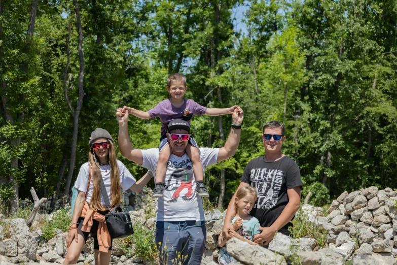 man with two children on his shoulders standing on a rock near a forest