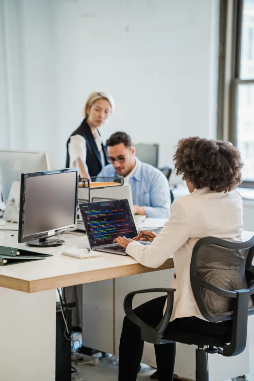 two people and one is sitting at a desk working on a computer