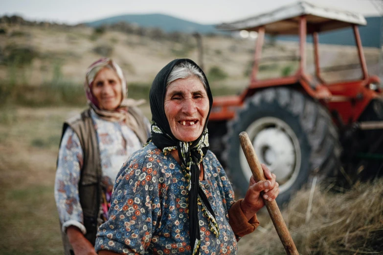 two women with large hair carrying a stick and a farmer's machine in the background