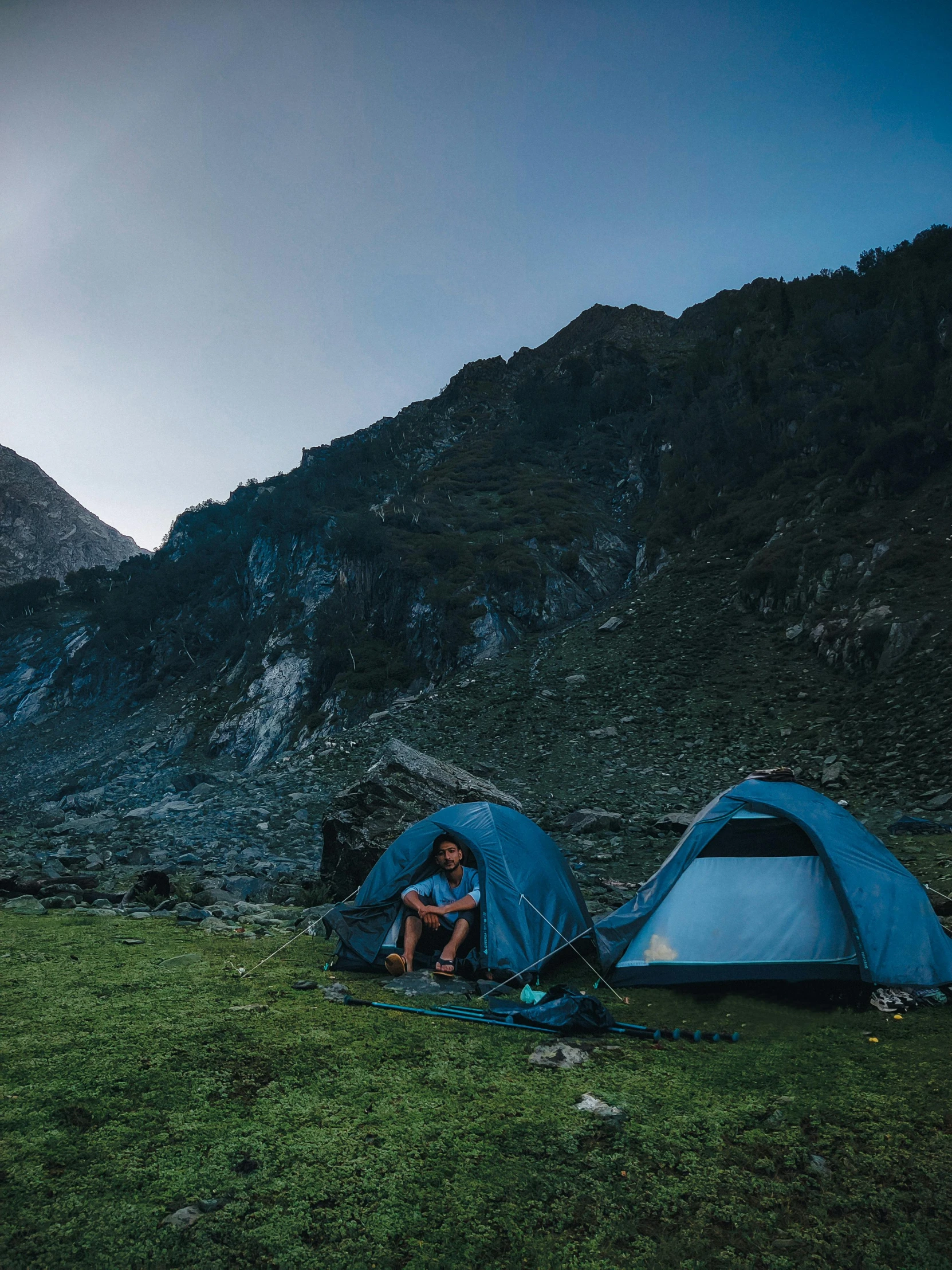 two blue tents sitting in front of mountains with a person kneeling next to one