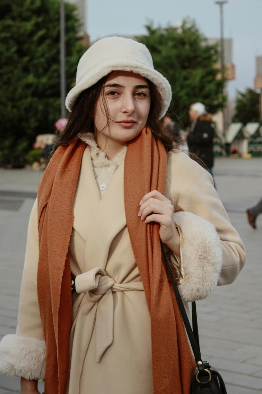 a young woman with a warm coat and orange scarf on her shoulders is wearing a winter hat and purse