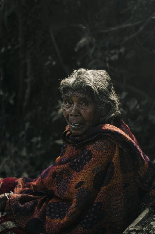 an older woman sits in a wooded area in her blanket