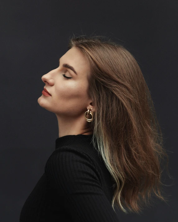 a woman with a dark background and a black background, she has her head tilted to the side she is wearing two yellow gold hoop earrings