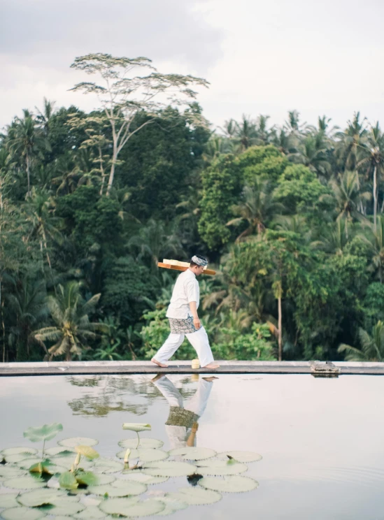 man in white walks beside pond with long stick on his head