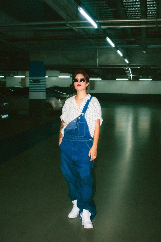 a woman with large sunglasses and overalls standing in an empty parking garage