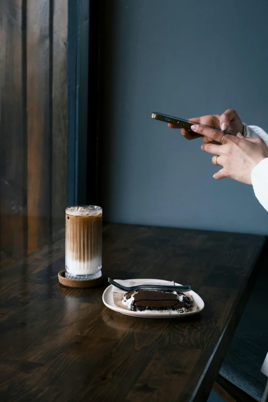 person holding a phone with two fingers and another hand next to a plate of chocolate cake