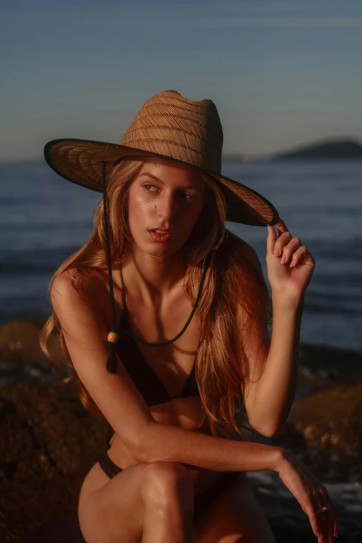 a woman is sitting down by the water with her hat on