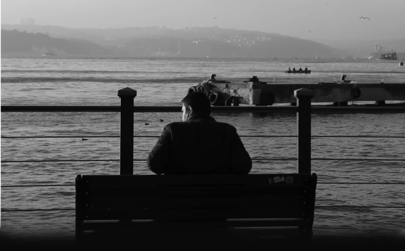 black and white po of a man sitting on a bench watching a boat pass by