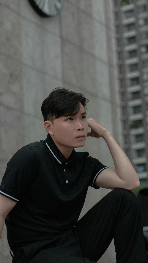 a young man in a black polo shirt and pants sits next to a stone building