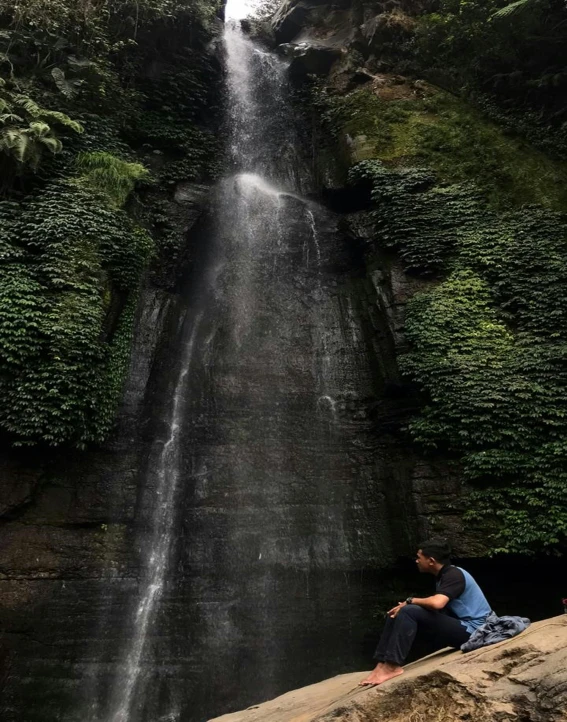 the man sits below the waterfall in front of the water