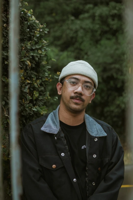 a young man with glasses and a cap stands in a park