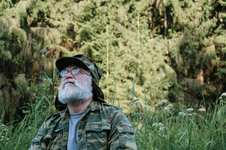 a man with a beard and glasses sitting in tall grass near trees