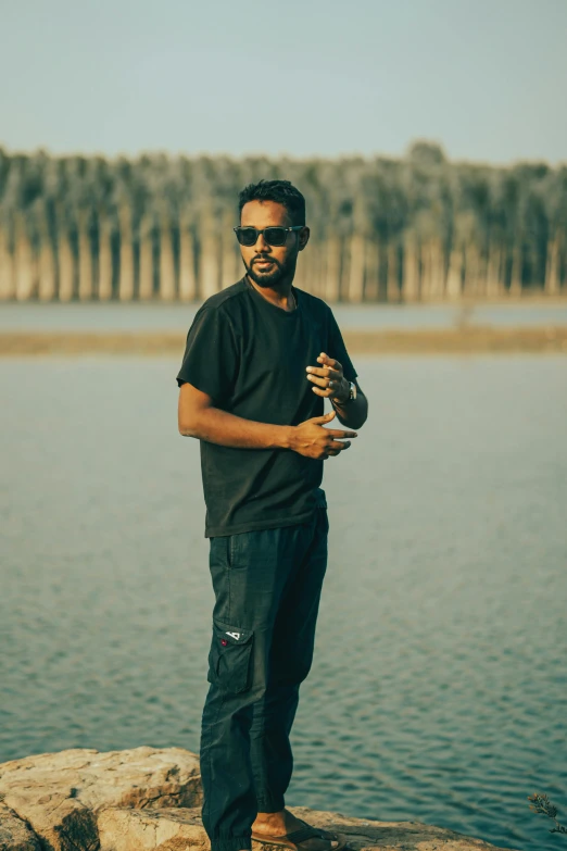 a man wearing sunglasses and holding a glass stands on a rock near water